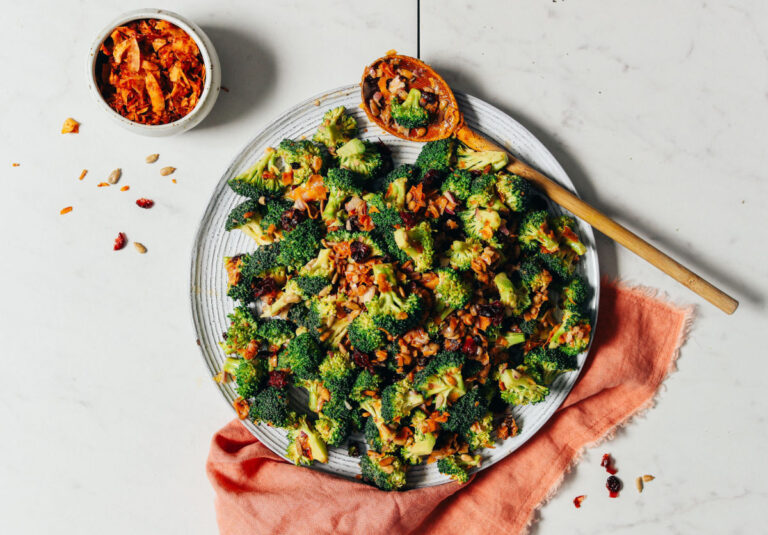 Image of broccoli salad topped with chilis on a plate, styled with a peach kitchen towel, a wooden spoon, and a small white bowl of toppings for this sulforaphane broccoli sprouts packed powerhouse salad.