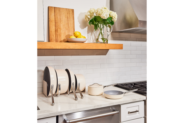 Image of beautiful kitchen with white marble countertop displaying Caraway Home Deluxe Set on the counter, with 4 pieces stored in their storage organizer. Above the countertop is a wood shelf with a glass pitcher of flowers, a wood cutting board, and a small dish of lemons.