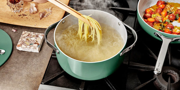 Image of Caraway Home Cookware pot in the color sage, with spaghetti being cooked, and pulled out with wooden tongs, on another burner on the range, is a saute pan with colorful small tomatoes cut in half with fresh herbs and olive oil. On the counter to the side sits the lid and a piece of garlic.