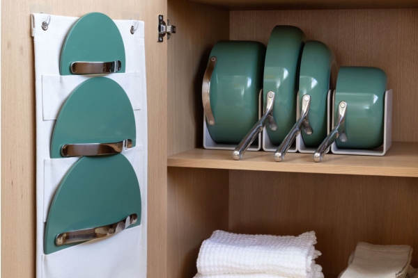Image of Caraway Home cookware set in sage, stored in cabinet with Caraway's storage organizers that comes with a canvas lid holder that hangs on the inside of the cabinet door and pot/pan holders that look great on a counter or in a wood cabinet, as they are pictured here. On the shelf below are white kitchen linens and towels. The Caraway cookware set comes with its own storage rack.  
