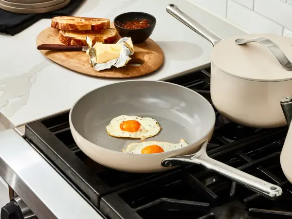 Image of the Caraway Home Frypan with two fried eggs on a modern range with a sauce pot and lid in the back burner, both in color cream. Next to the range on a white marble counter sits a small oval wood cutting board with toast and butter.