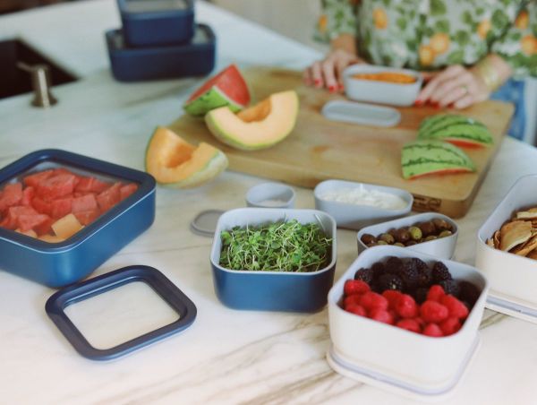 Image of a food prep session displayed on a white marble kitchen island with wood cutting board. Caraway Home food storage containers (in white and blue) filled with fresh berries, cut melon, olives, and microgreens.