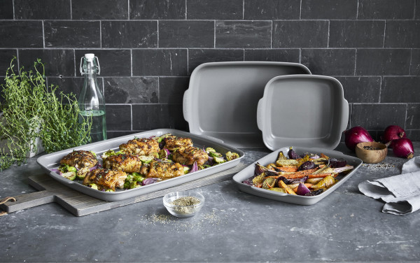 Image of Greenpan premiere nontoxic bakeware set on grey concrete counter with dark slate backsplash. One GreenPan baking sheet is on a cutting board with roasted chicken breasts with broccoli, and another pan sits next to it with roasted root veg and onion.