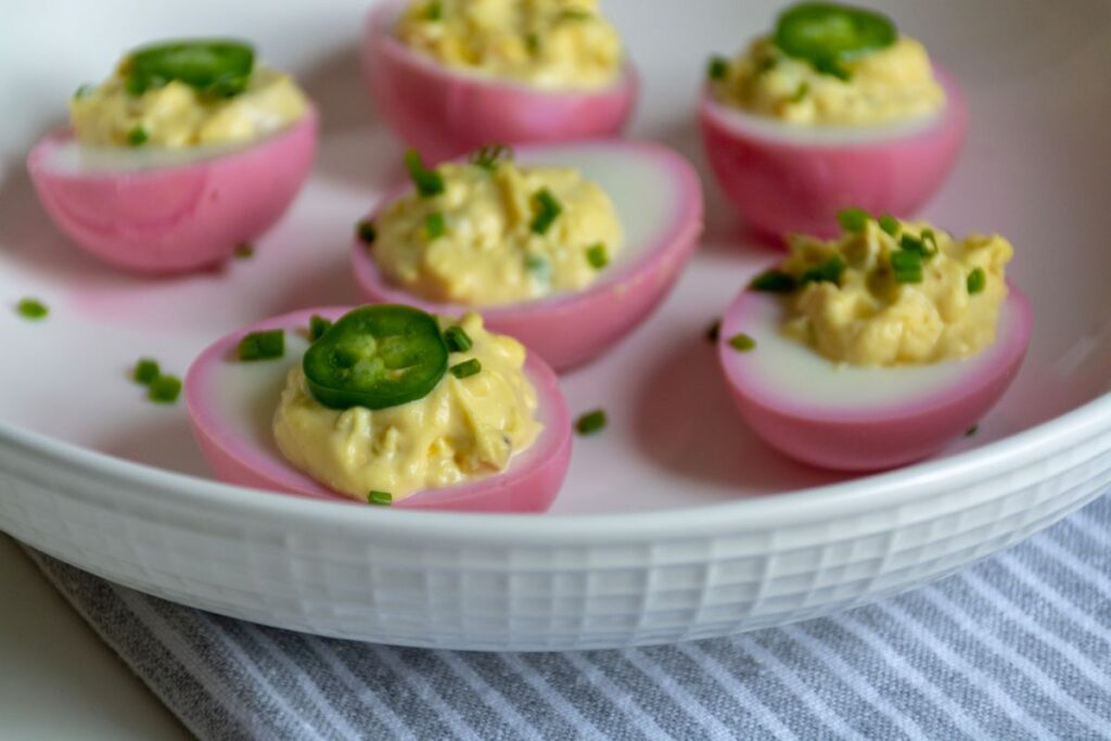 Image of pink beet pickled deviled eggs on a white plate on top of a blue stripped kitchen towel. Serve these for Easter and add a pop of color to your spread.