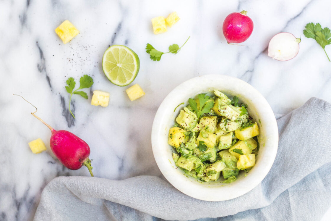 Image of a white bowl of cubed pineapple with cilantro on gray and white marble surface with bright radishes and a slice of lime showing how to overcome eating unhealthy food and change your health.
