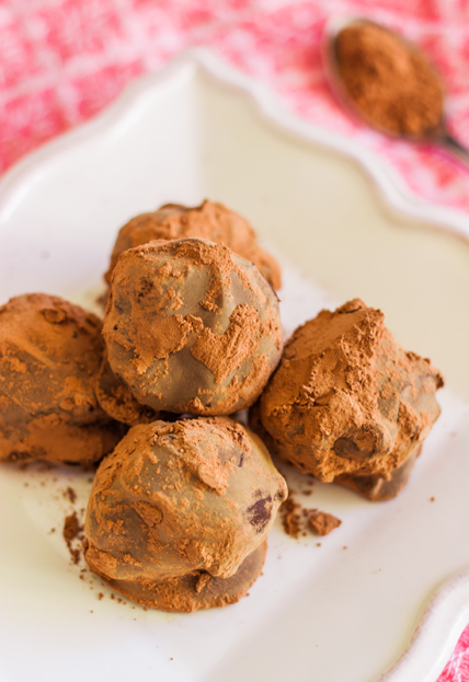 image of 5-ingredient chocolate truffle recipe, small chocolate truffle balls with pure cacao dusted on top, in a white serving dish on red checked tablecloth blurry in the background. This easy chocolate truffle recipe comes together in no time and is perfect for gift-giving.