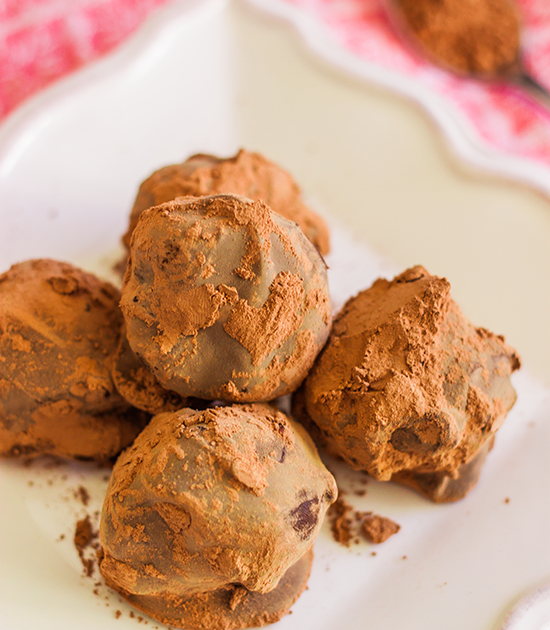 image of 5-ingredient chocolate truffle recipe, small chocolate truffle balls with pure cacao dusted on top, in a white serving dish on red checked tablecloth blurry in the background. This easy chocolate truffle recipe comes together in no time and is perfect for gift-giving.