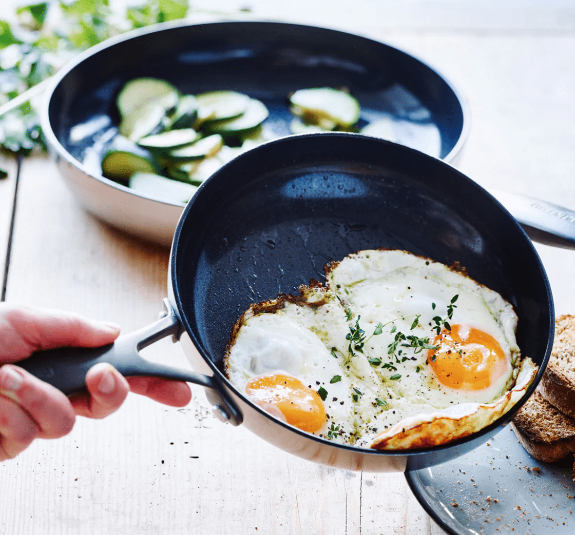Image of two Venice Pro Noir frypans, the pan in the forefront has fried eggs being easily slid out with herbs and the pan in the background has fresh veggies, off to the side there is a wood cutting board with herbs and seasonings and tongs. You’d be very happy if you bought this frypan set, with its matte black modern handles. Search for the line you want, select the set you want, add to cart is easy, and bring these to a happy kitchen.