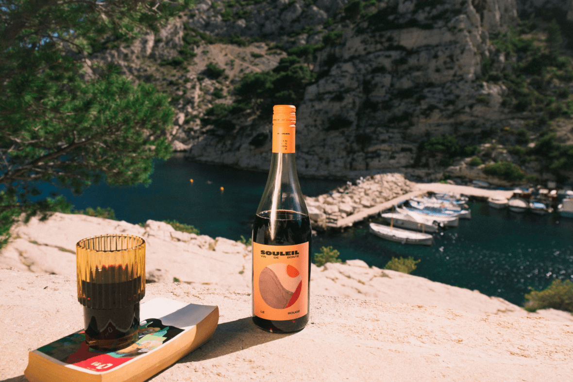 Image of a bottle of Souleil wine on a rocky cliff overlooking deep emerald water and boats.