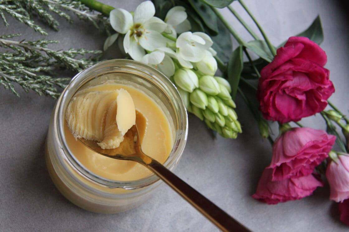 image of an overhead shot of a small glass pot of ghee with a silver spoon scooping it out for Ayurvedic cleanse. There are fresh pink and white flowers next to the ghee on a white cloth.
