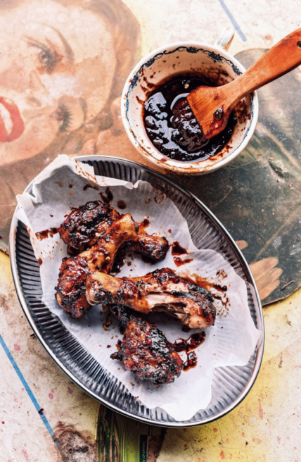 Image: messy, saucy barbecue chicken on white parchment paper on top of a black plate with small white bowl of extra sauce on the side with wooden sauce brush all sitting on a well-worn colorful table. Grilled bbq chicken recipe is saucy and flavorful via Organic Authority.
