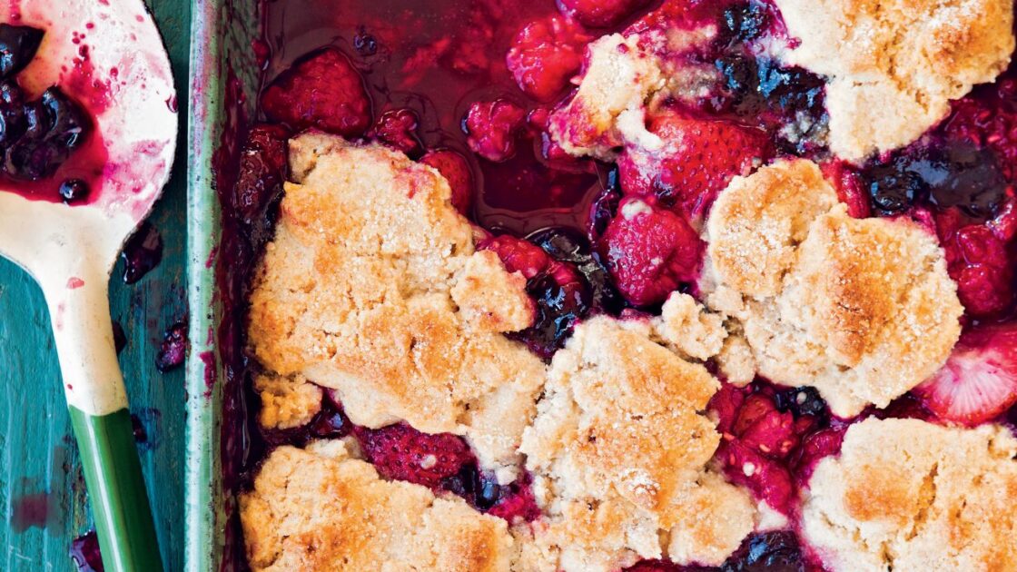 Image: berry cobbler with a deep pink filling blueberries, raspberries, strawberries, and a biscuit like topping in a turquoise ceramic baking dish on a worn turquoise wooden table, serving spoon set to the side. This easy berry cobbler recipe comes together so easily via Organic Authority.