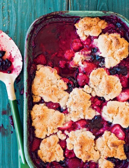 Image: berry cobbler with a deep pink filling blueberries, raspberries, strawberries, and a biscuit like topping in a turquoise ceramic baking dish on a worn turquoise wooden table, serving spoon set to the side. This easy berry cobbler recipe comes together so easily via Organic Authority.