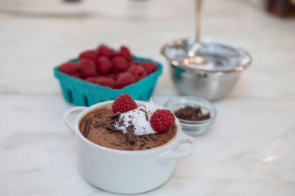 Image of blender chocolate mousse served in a white ramekin with small handles, topped with whipped cream and fresh raspberries. A small pint of raspberries sit in teal container in the background, along with shaved chocolate in a small glass ramekin.