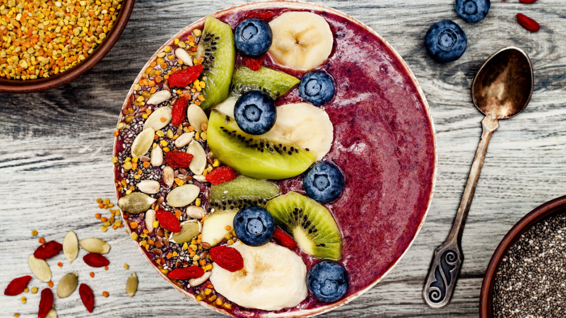 Image of a colorful smoothie bowl for a healthy breakfast with sliced kiwi, and bananas, fresh blueberries, dried goji berries, chia seeds and pumpkin seeds on a worn wooden surface with an aged silver spoon and extra ingredients sprinkled around.