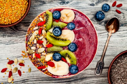 Image of a colorful smoothie bowl for a healthy breakfast with sliced kiwi, and bananas, fresh blueberries, dried goji berries, chia seeds and pumpkin seeds on a worn wooden surface with an aged silver spoon and extra ingredients sprinkled around.