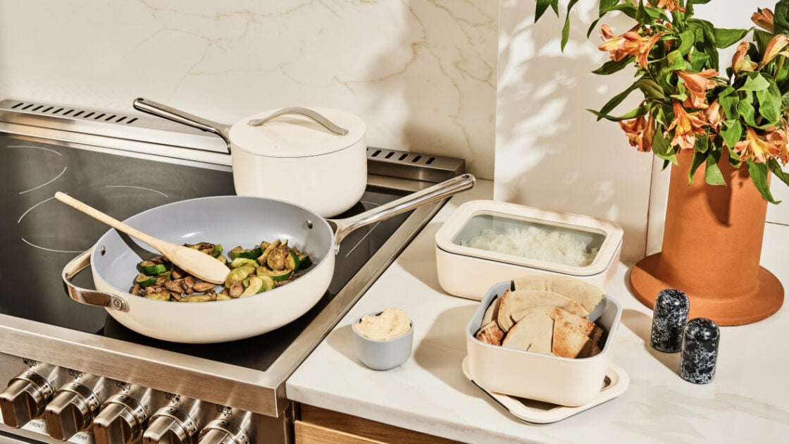 Image of Caraway Food Storage in cream, on a white marble countertop, prepped alongside a flat stovetop with amCaraway Home cookware Fry Pan and a covered saucepan to the rear. There's also a terracotta vase with fresh lilies, black marble salt and pepper shakers, and a veined marble backsplash.