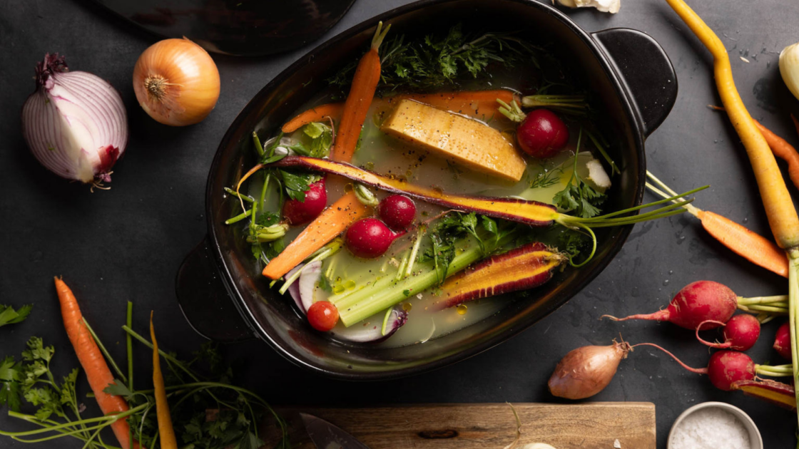 Image of Xtrema ceramic dutch oven on countertop filled with broth and fresh cut vegetables to make soup. On the countertop, there are more fresh veggies, fresh quartered red onion, shallots, radishes and carrots.