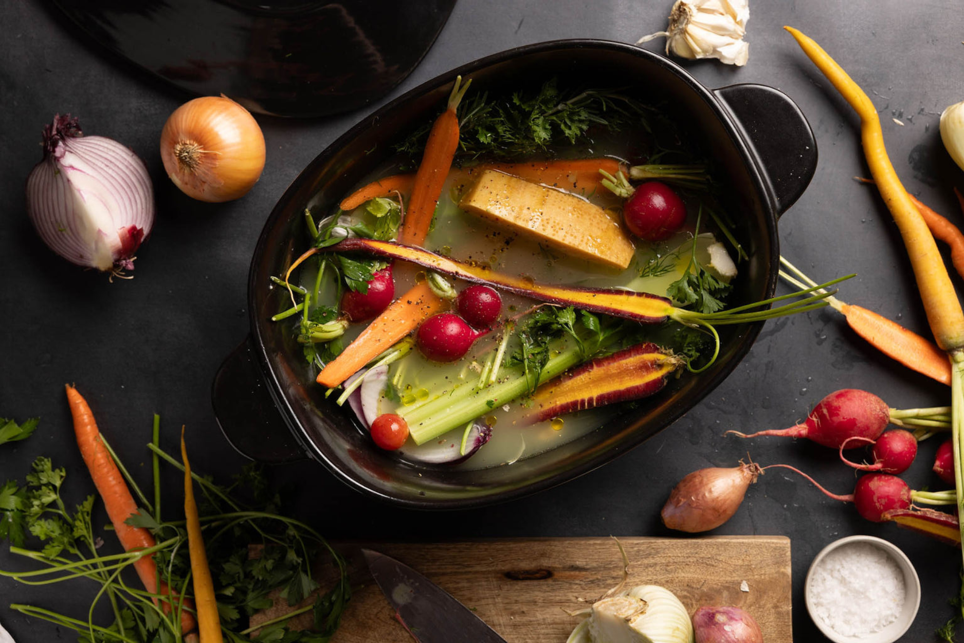 Image of Xtrema ceramic dutch oven on countertop filled with broth and fresh cut vegetables to make soup. On the countertop, there are more fresh veggies, fresh quartered red onion, shallots, radishes and carrots.