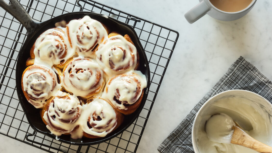 image of a Homemade vegan cinnamon roll recipe or buns baked in a cast iron skillet and covered with cream cheese icing. The skillet is cooling atop a black wire cooling rack on a white and gray marble countertop. A mug of coffee or tea is on the side next to a bowl full of icing.