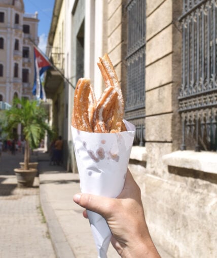 Image of a woman's hand holding a paper cone filled with sugar-covered churros on the streets of Cuba. This easy churros recipe is one you can make at home and it happens to be vegan.