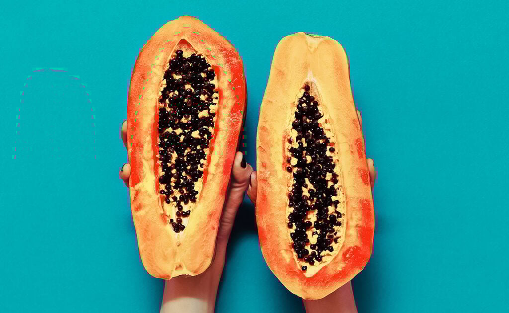 Image of cut open papaya held up on turquoise wall by two hands. Papaya is an excellent for supporting digestive enzymes