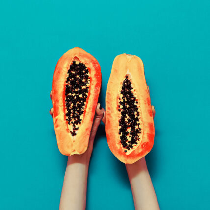 Image of cut open papaya held up on turquoise wall by two hands. Papaya is an excellent for supporting digestive enzymes