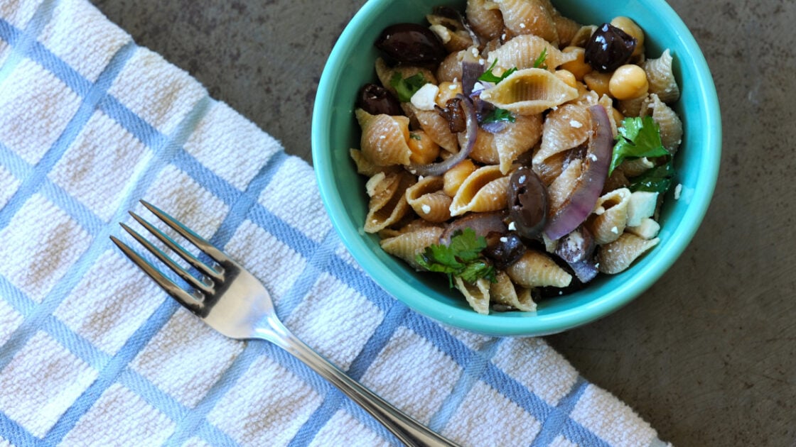 Image of a small blue bowl filled with pasta salad featuring feta and chickpeas, next to a napkin and fork.