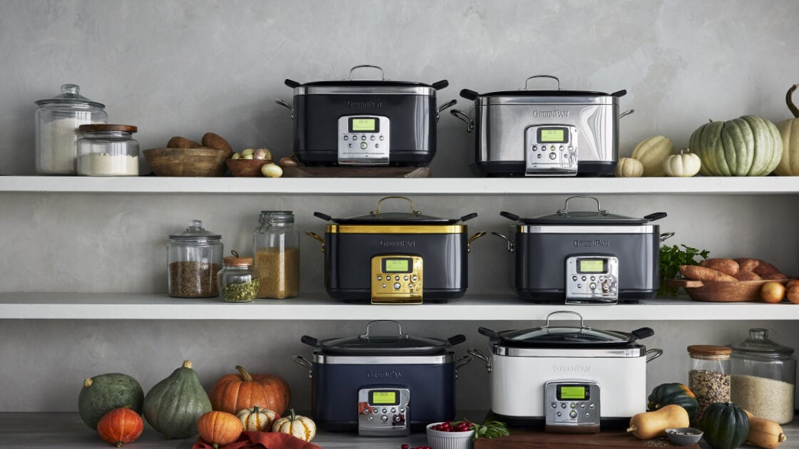 Image of 6 GreenPan Slow Cookers in various colors/finishes/ and lines. Lined up on the counter and two shelves above. The image is also styled with squash and pumpkins, glass canisters of pantry staples, and a wood cutting board.