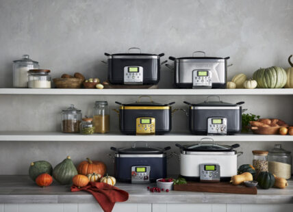 Image of 6 GreenPan Slow Cookers in various colors/finishes/ and lines. Lined up on the counter and two shelves above. The image is also styled with squash and pumpkins, glass canisters of pantry staples, and a wood cutting board.