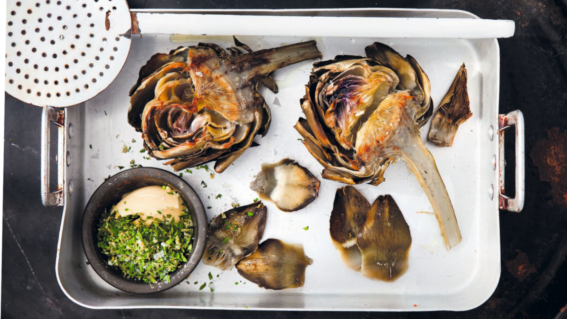 Image of grilled artichokes recipe in a white roasting pan with a black background and a small black ramekin of gremolata nestled in the corner, a white spider (large slotted spoon) rests on top of the roasting pan.