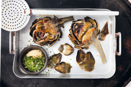 Image of grilled artichokes recipe in a white roasting pan with a black background and a small black ramekin of gremolata nestled in the corner, a white spider (large slotted spoon) rests on top of the roasting pan.