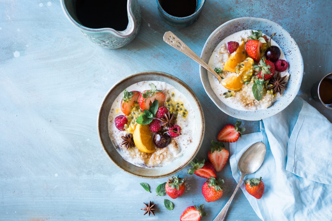 Image of two bowls of oatmeal as a healthy breakfast option covered in mixed fresh cut fruit on a concrete worktop, with fresh strawberries strewn about and a light blue tea towel and a side of maple syrup.