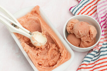 Image: Pink watermelon sorbet or ice cream scooped into a white bowl with white ice cream scoop out of a white enamel bread pan with colorful striped towel off to right side via Organic Authority.
