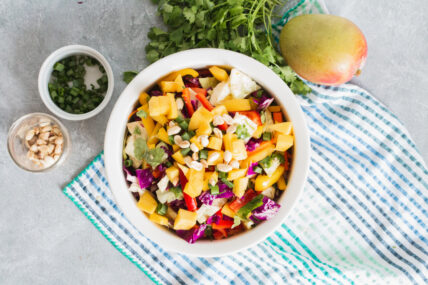 Image: mango salad in a white bowl surrounded by peanuts green onions, cilantro and a whole mango on a striped blue and white towel via Organic Authority.