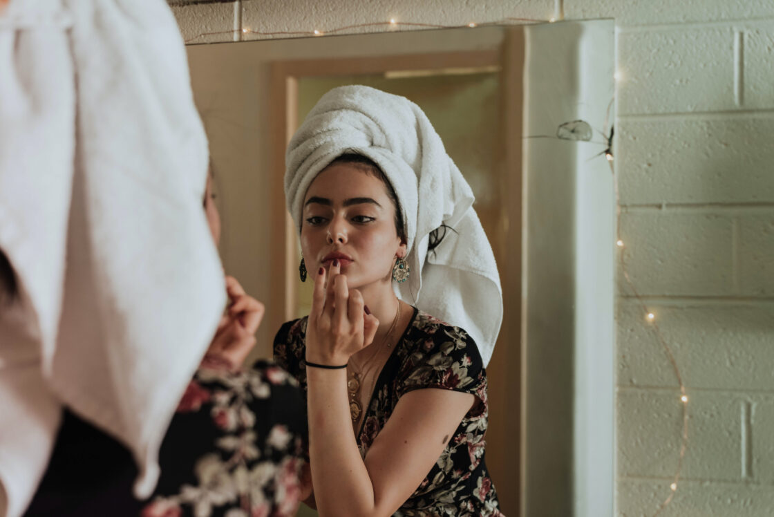 Cortisol is a hormone that affects women and can trigger hormone imbalance symptoms - Woman applies makeup and skincare in mirror with towel on her head.