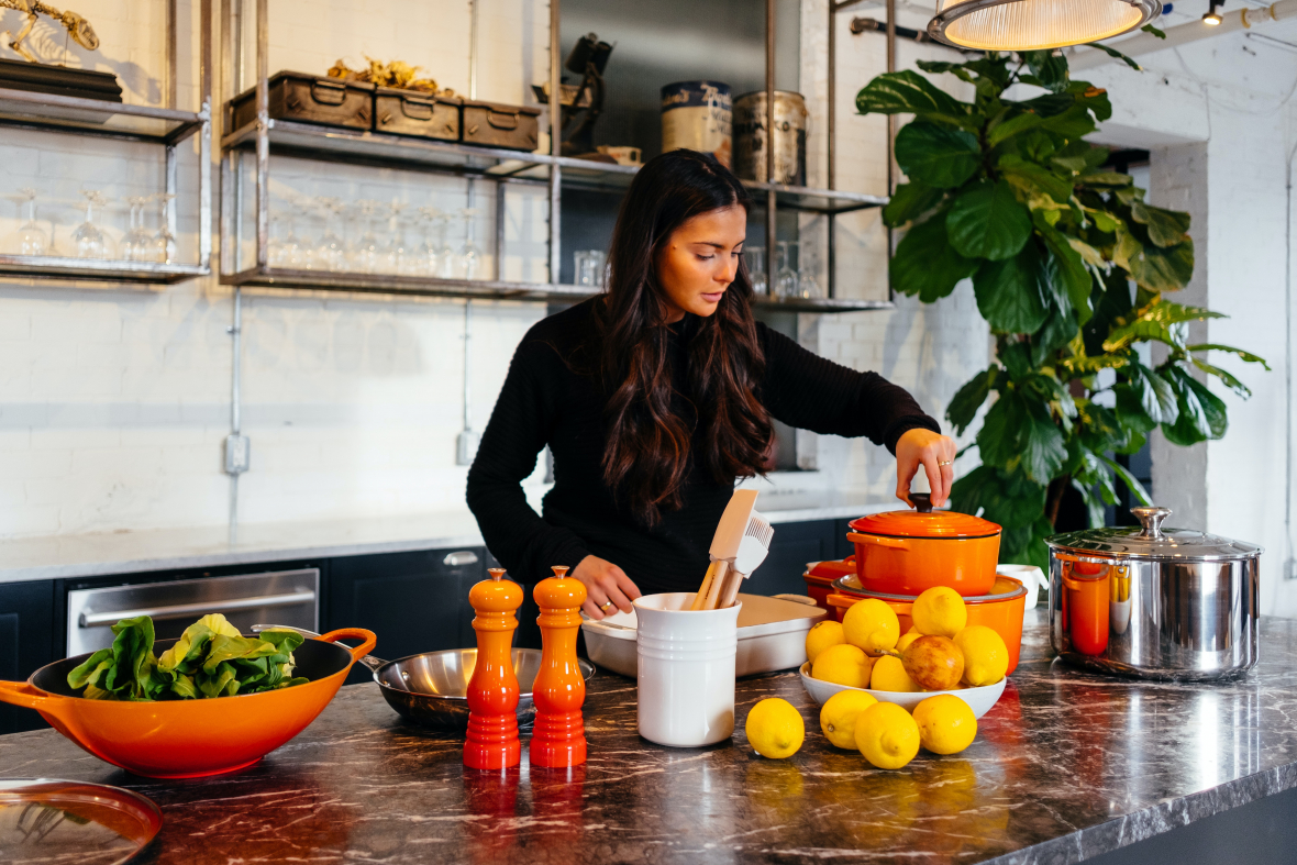 image of long brown-haired woman setting lid on bright orange pot stacked on larger orange pot at marble island with bowl of lemons and pan of greens which are both liver detox ingredients.