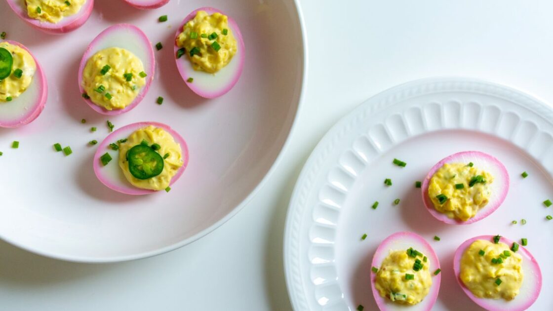 Image of pickled deviled eggs on two white plates. The eggs are pretty with a pink exterior, giving a lovely pink rim around the cut side, filled with yellow yolk filling and topped with chives and thinly sliced jalapenos.
