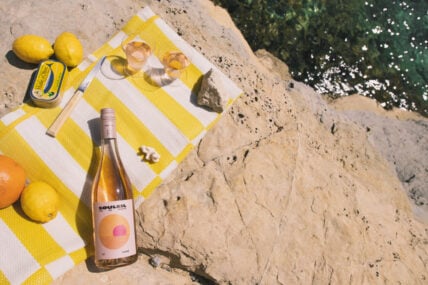 Image: Bottle of summer wine, Souleil, displayed on a bright yellow and white striped towel, with 2 glasses of light pink wine served in small glasses, lemons and an orange, a can of sardines, and small knife all on rocky cliff with small peek of sparkling ocean below.