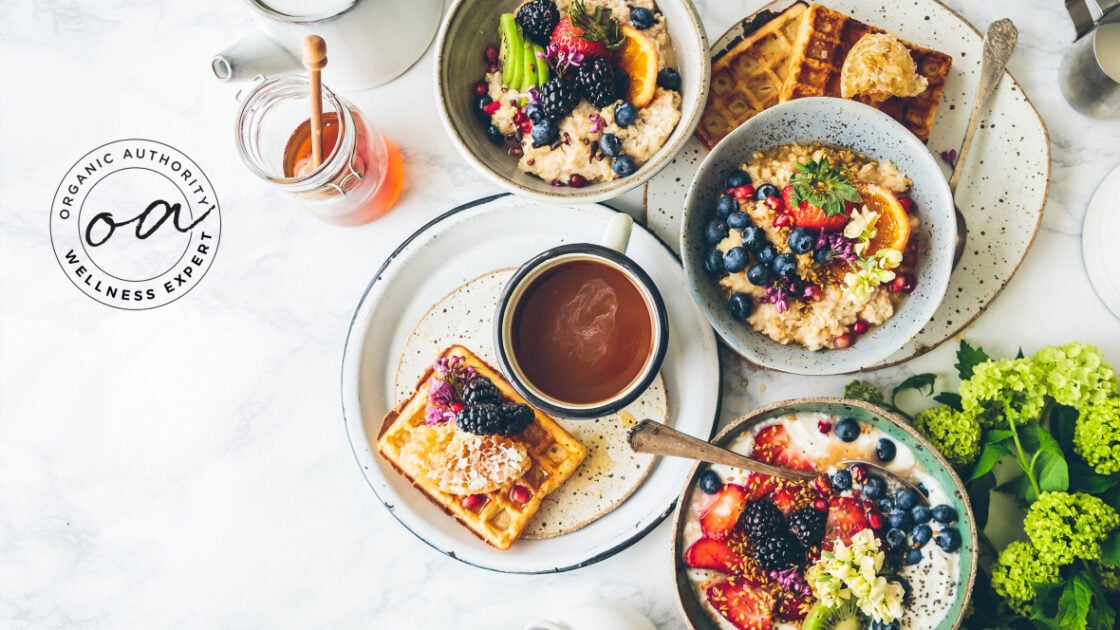 Image of an overhead shot of a mix of unhealthy food, poor nutrition, and healthy food, all plated on speckled white plates on a white marble surface. Waffles, oatmeal, berries, and a jar of honey with Organic Authority Wellness Expert seal, demonstrating the link between diet and disease.