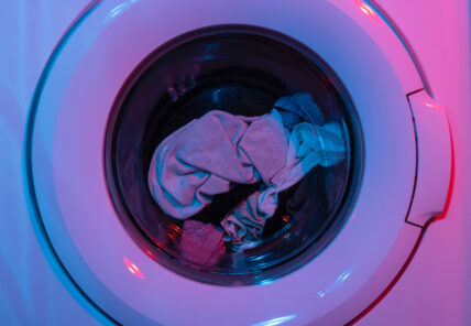 Image of front loading washer with purple and pink lighting, giving a cool yet eerie look to show that toxic chemicals in laundry detergent. Dry clothing is inside of the clear glass front washer door.