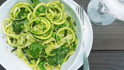 Image of spiralized zucchini pasta recipe with Tagliatelle, Peas, and Arugula Pesto, served in white pasta bowl, with charger beneath and a fork. Served on wood table with wine glass.