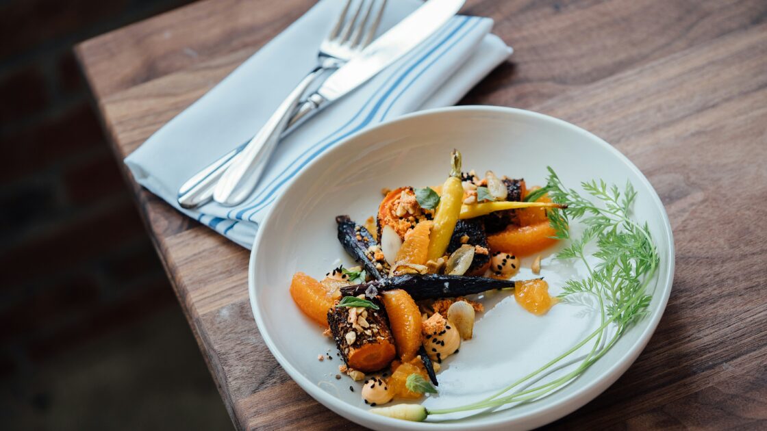 Image of a plate of carrots and orange segments artfully plated on a wood table with a folded cloth napkin with silverware. Least Expensive Fruits and Veggies, Fruits and Vegetables, Unsplash