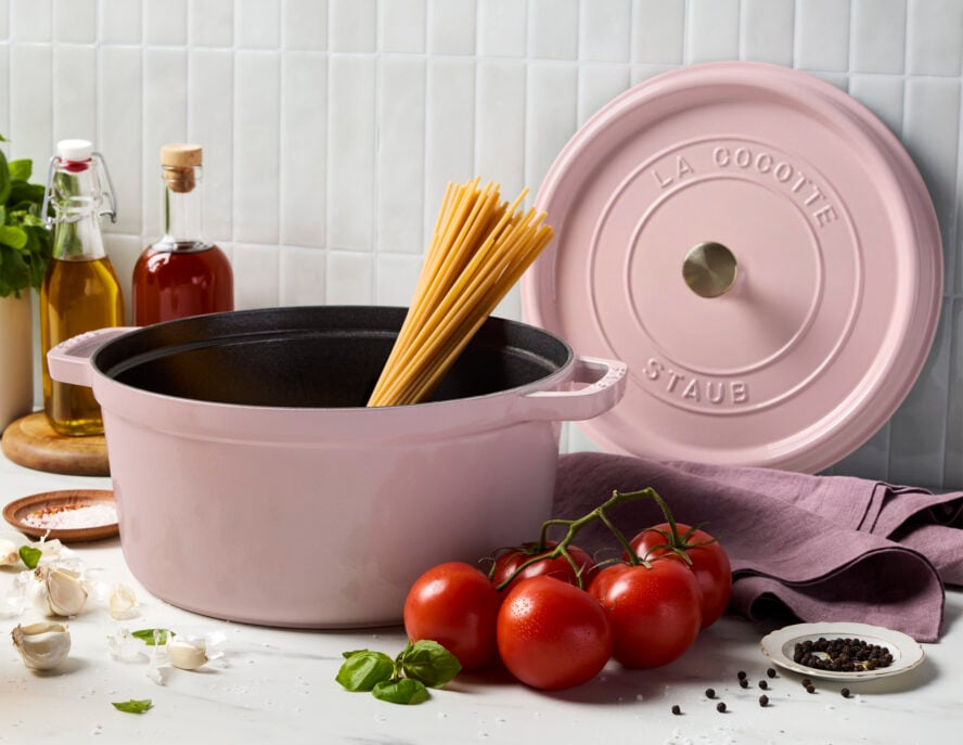 Image of the Staub round cocotte in Pink Peony, filled with uncooked spaghetti noodles, with a lid leaning against the backsplash on the wall.