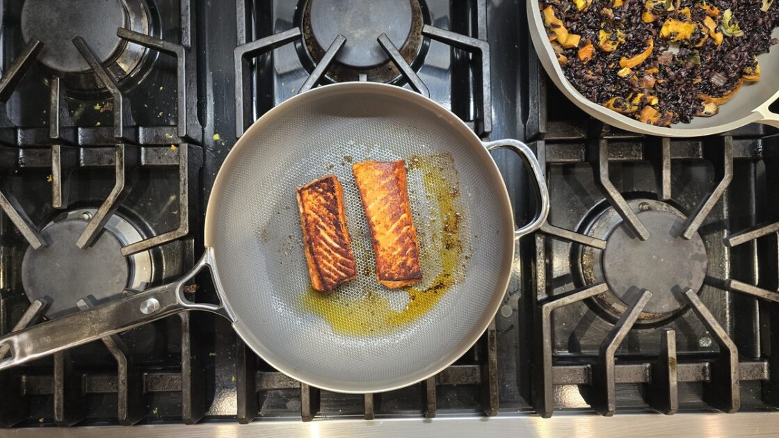 Image of a gas stovetop with two Our Place fry pans on it, one cooking two salmon filets.