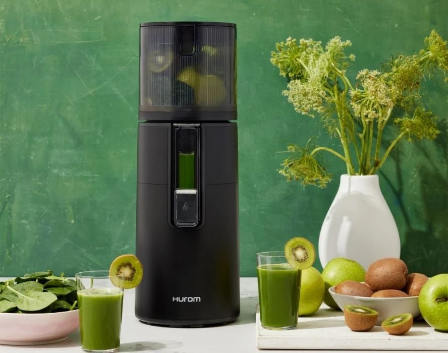 One of the many kitchen gifts recommended by the OA Editor-in-Chief, the Hurom H400 Easy Clean Slow Juicer. It's displayed on a white table with a green background, surrounded by kiwis, green apples, and leafy greens.