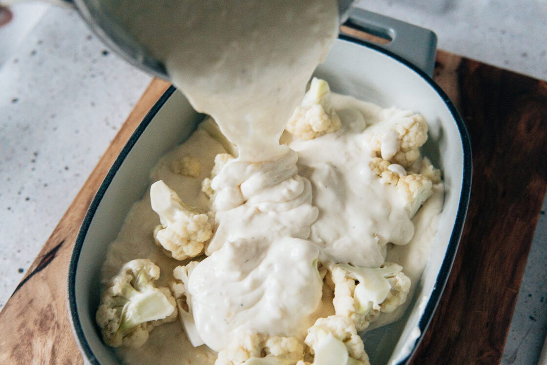 Image of cheesy roux being poured atop a layer of cauliflower florets in a Pampered Chef Enameled Cast Iron Baker for maximum cheesiness.
