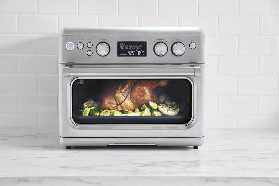 Image of a silver GreenPan Elite Convection Air Fry Oven on a countertop roasting a whole chicken and green vegetables.