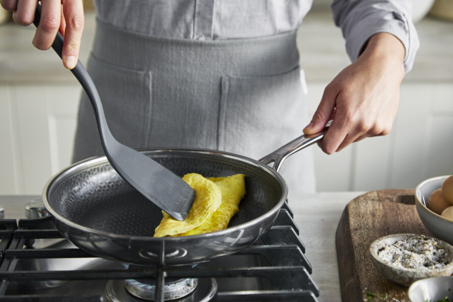 Image of a pair of hands holding a GreenPan Spectra frypan and frying an omelette with a spatula on the stovetop.