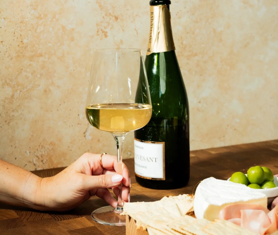 Image of someone's hand holding a Made In Crystal Wine Glass, filled with white wine. To the right is a wooden serving board with crackers, cheese, and olives.